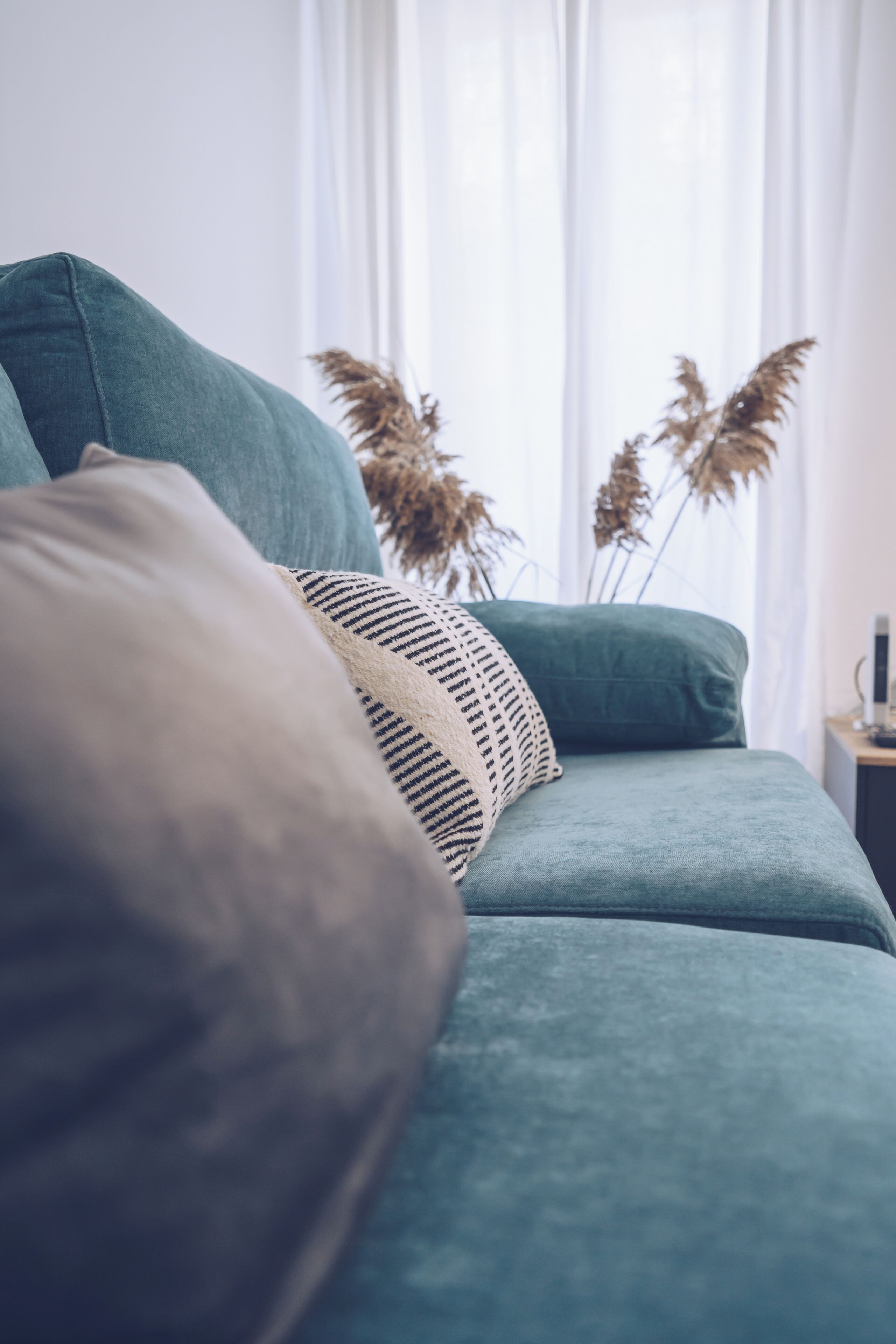 A teal sofa with a patterned pillow, a vase with dried plants, and a small table with a lamp.