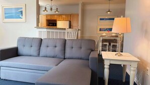 A modern hotel room with a grey sectional sofa, a small white table with a lamp, a framed picture on the wall, and a view of the kitchen area. - Runaway Beach Club 1 Bd Villa (LRW 17101) (Kissimmee)