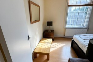 A bedroom with a bed, a bedside table with a lamp, a window with blinds, and a door. - Stockwell Central Apartment C (London)