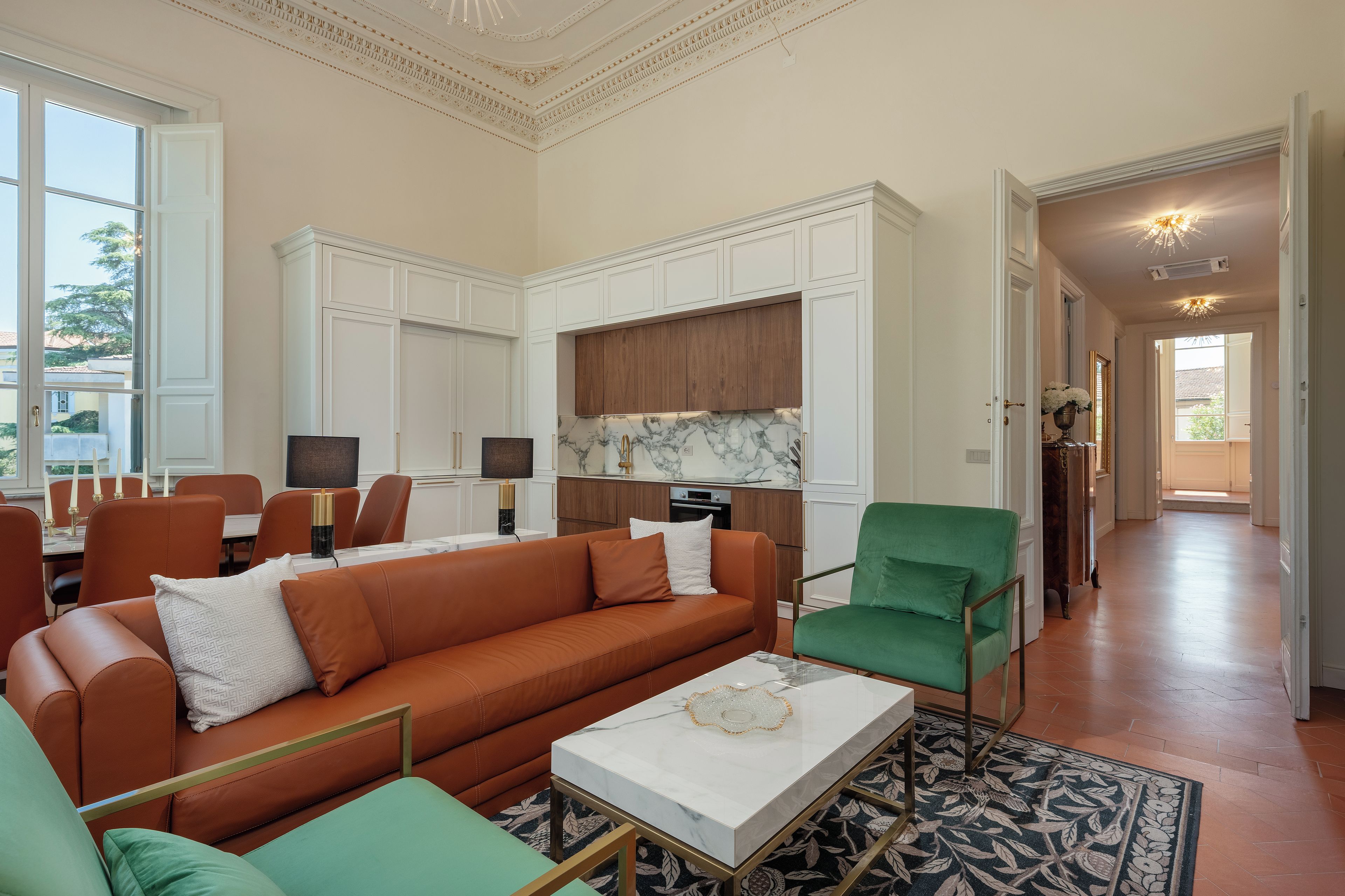 Een ruime woonkamer met een bruine leren bank, een groene fauteuil en een witte salontafel. Er is een keuken met witte kastjes en een marmeren achterwand.