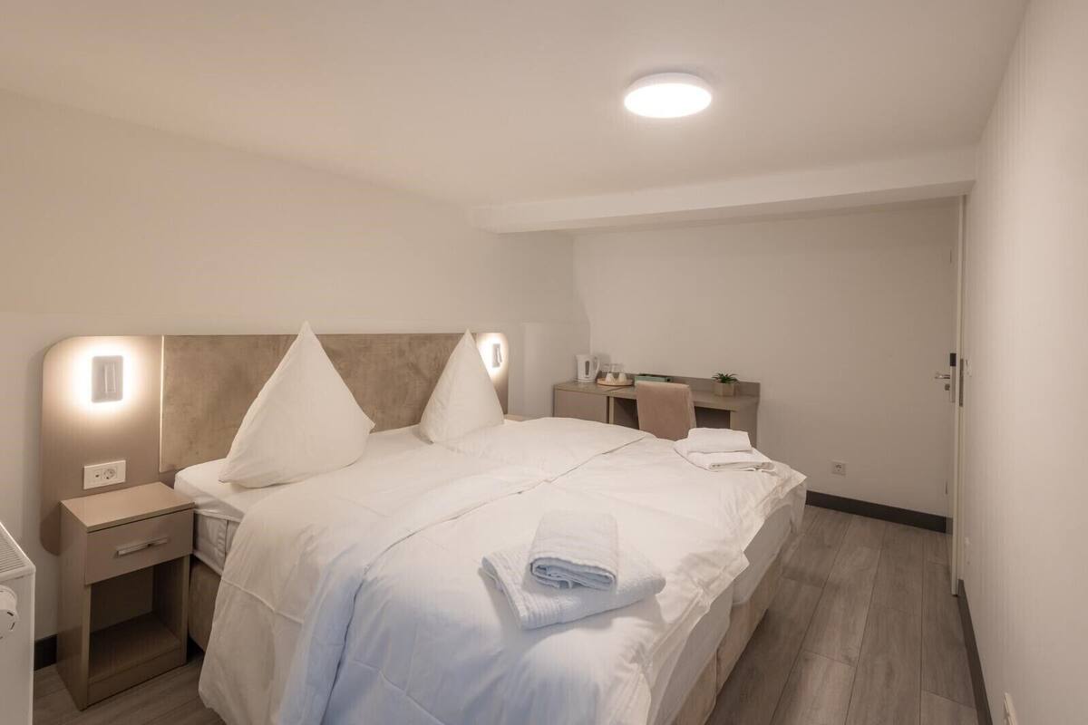 A modern hotel room with a neatly made bed, bedside tables, a desk with a chair, and a small plant.