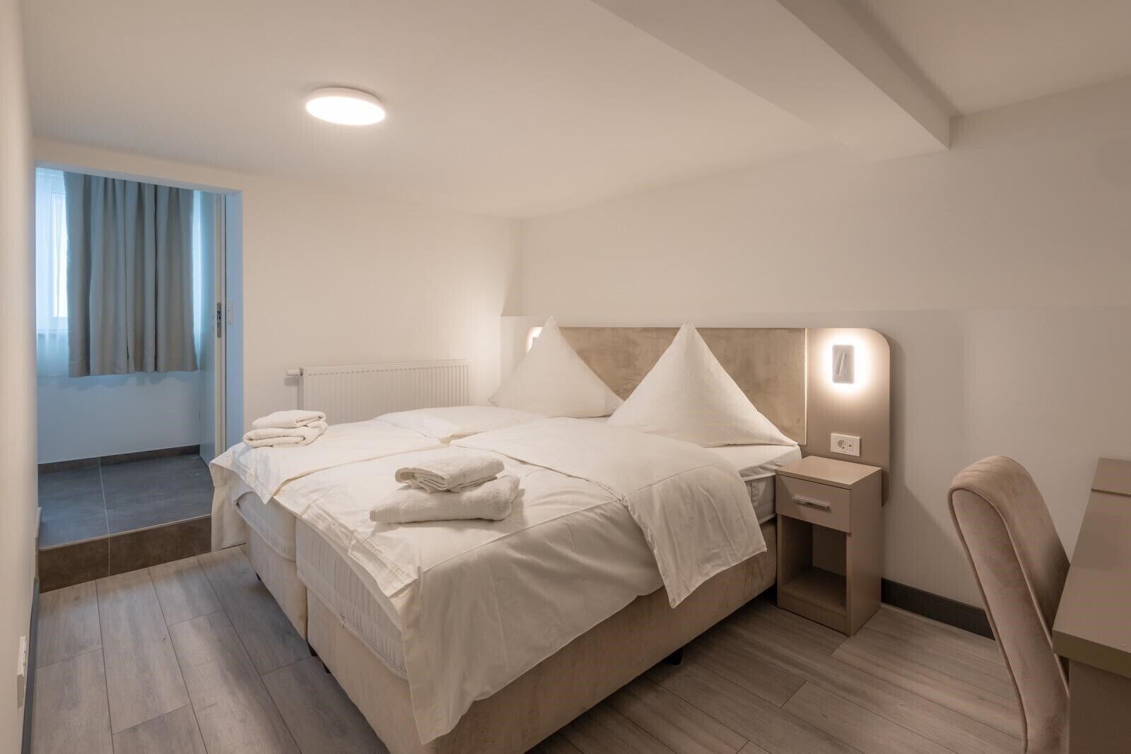 A modern hotel room with a large bed, a bedside table, a chair, and a window with curtains.