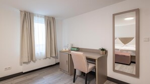 A modern hotel room with a wooden floor, a desk with a chair, a large mirror, a radiator, and a bed with a headboard. - Hotel Johannisberg (Bad Homburg v.d. Hoehe)