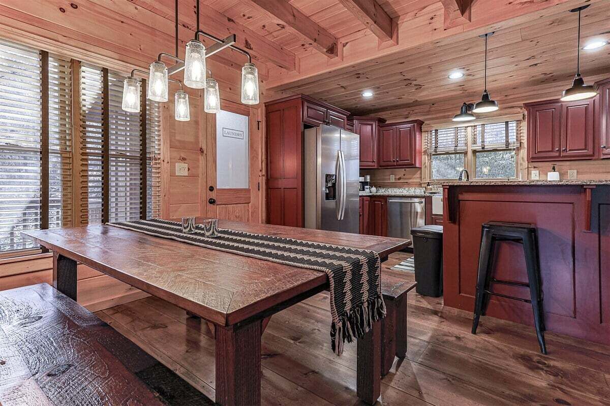 A wooden kitchen with a large dining table, hanging pendant lights, and a refrigerator.