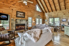 A log cabin interior with a stone fireplace, wooden ceiling beams, a sofa, a dining table, and a kitchen area. - Heavenly View Lodge | Ellijay, GA (Ellijay)