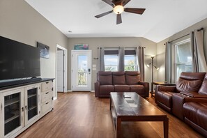 A living room with a flat-screen TV, leather sofa, wooden coffee table, ceiling fan, and a view of the outdoors. - Bay-View Delight / Sleeps 8 (Rockport)