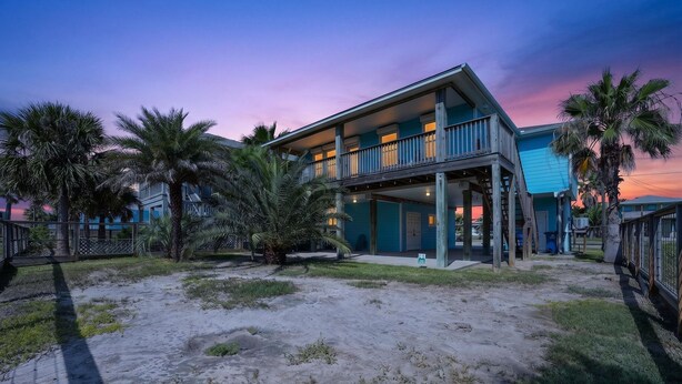 Een tweelaags pand aan het strand met een balkon, palmbomen en zand.