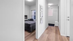 Un couloir mène à une chambre qui contient un lit, une table de chevet et une fenêtre. Des tableaux encadrés ornent le mur, et une porte se trouve sur le côté droit.