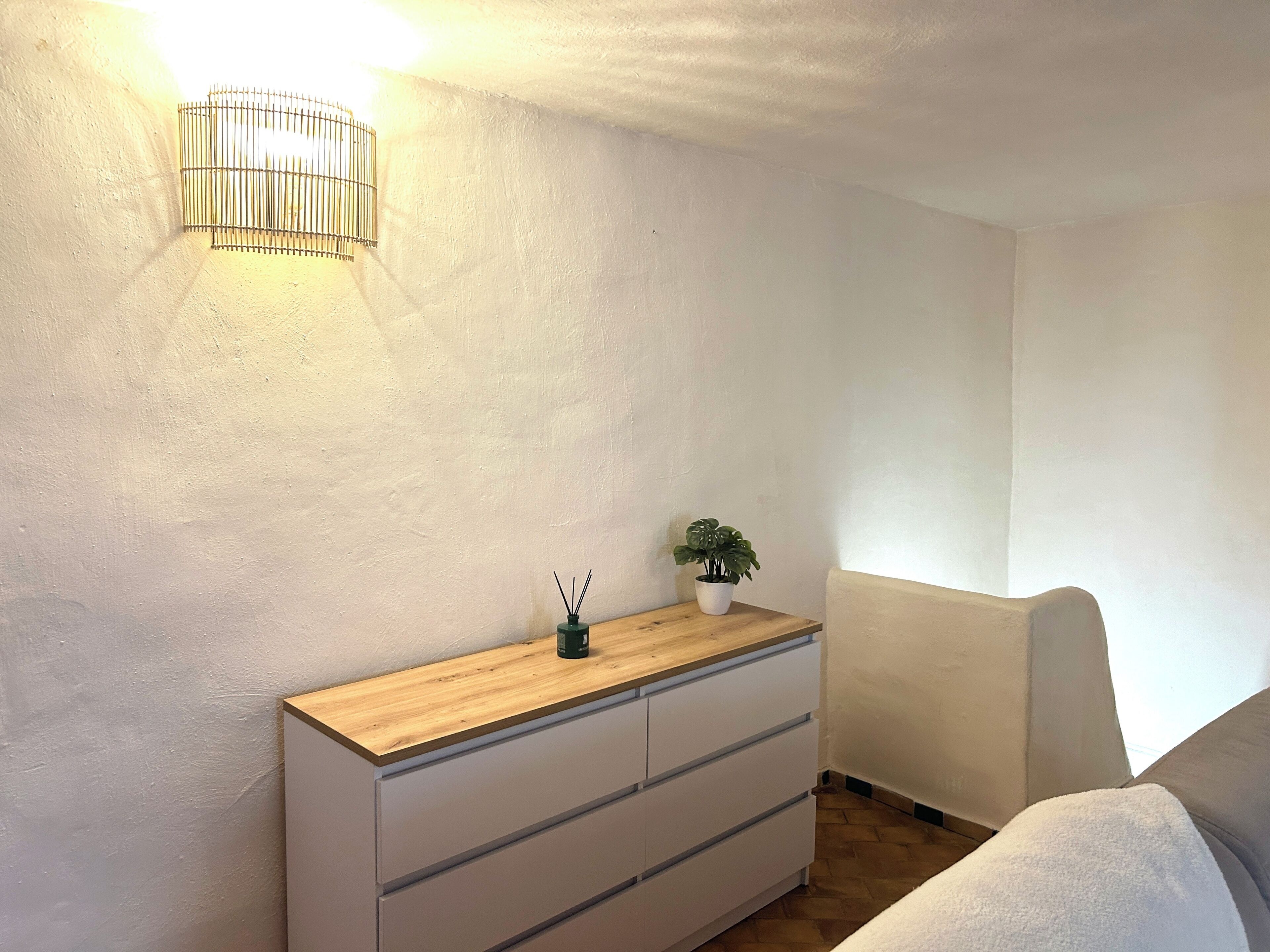 Quarto com cómoda de madeira, uma pequena planta em vaso e luminária fixada na parede.