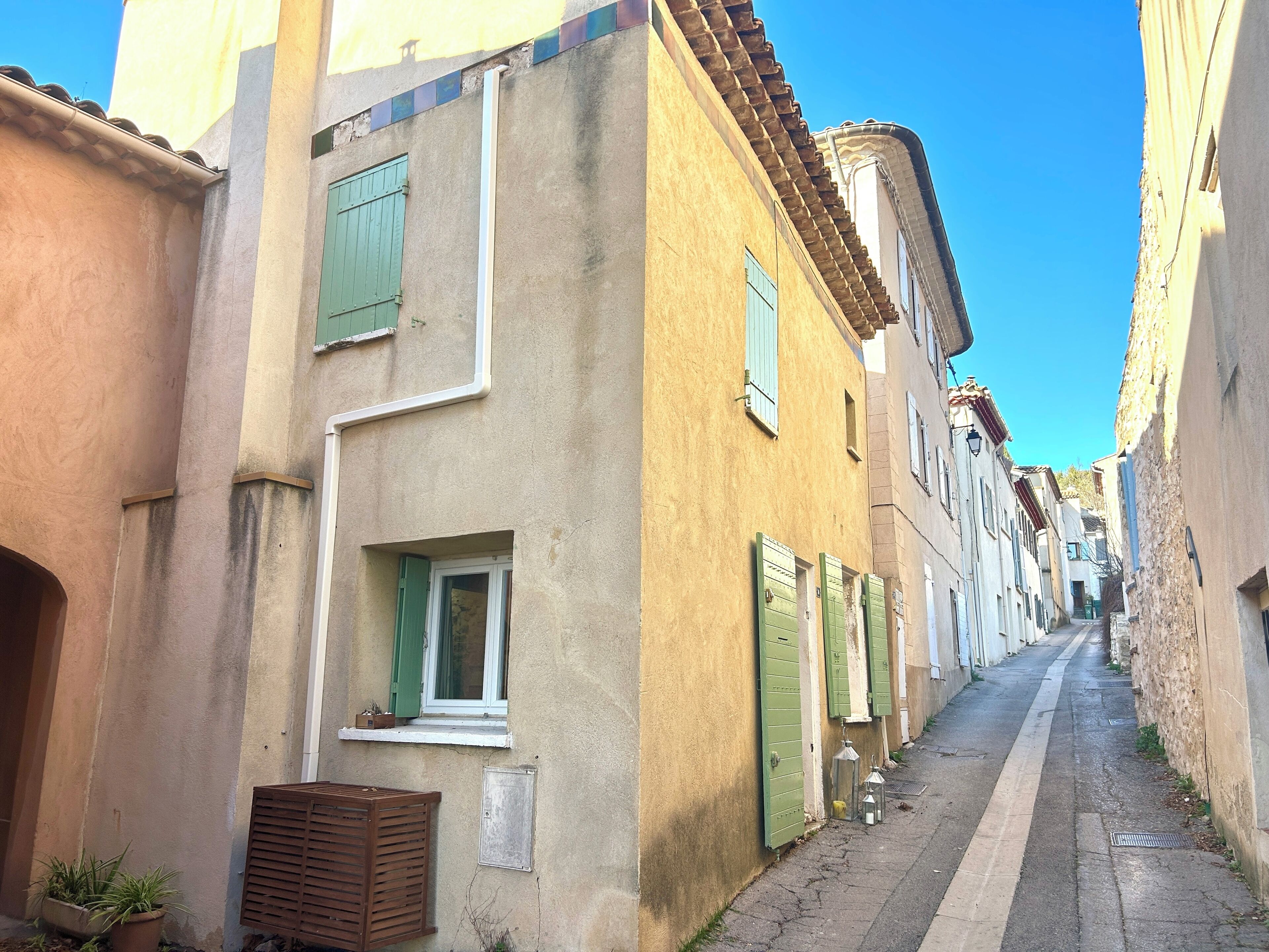 A narrow street lined with traditional buildings, featuring small windows with shutters, a visible door, and a small balcony with a potted plant.