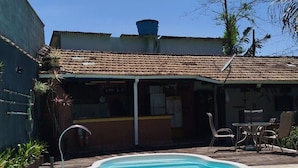 Una piscina amb un solàrium d'rajola, una zona de pati coberta amb mobles d'exterior i un edifici amb teulada enrajolada al fons.