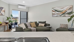Un salon moderne avec un ensemble de canapés gris, une table basse et un coin repas agrémenté d’un buffet en bois. Deux grandes peintures abstraites ornent les murs, et une grande fenêtre offre une vue sur la ville.