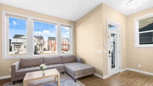 A living room with a sectional sofa, a coffee table, and a view of a residential neighborhood through large windows. - Family-Friendly 3-Bed | Super Close to Airport! (Calgary)