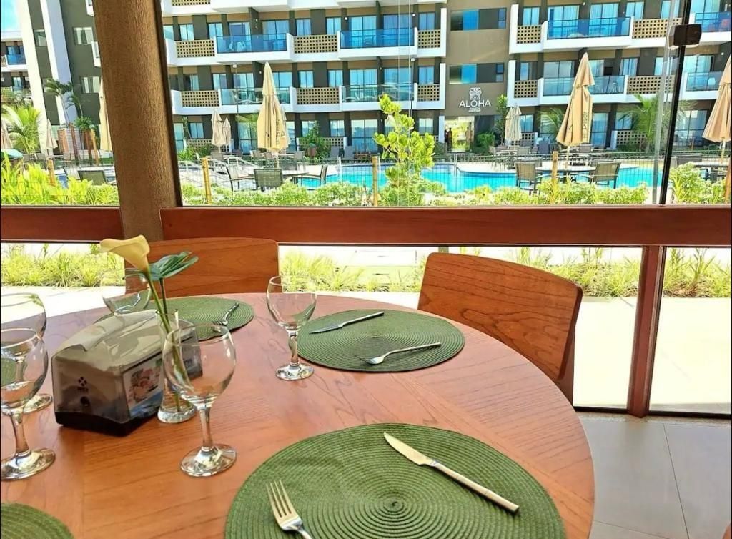 A dining table set with green placemats, cutlery, and a glass. A vase with a yellow flower is on the table. The background features a pool and a hotel building with balconies.