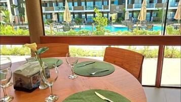 A dining table set with green placemats, cutlery, and a glass. A vase with a yellow flower is on the table. The background features a pool and a hotel building with balconies.