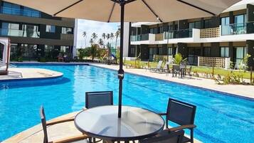Zona de piscina con mesa y sillas, una gran sombrilla y un edificio moderno al fondo.