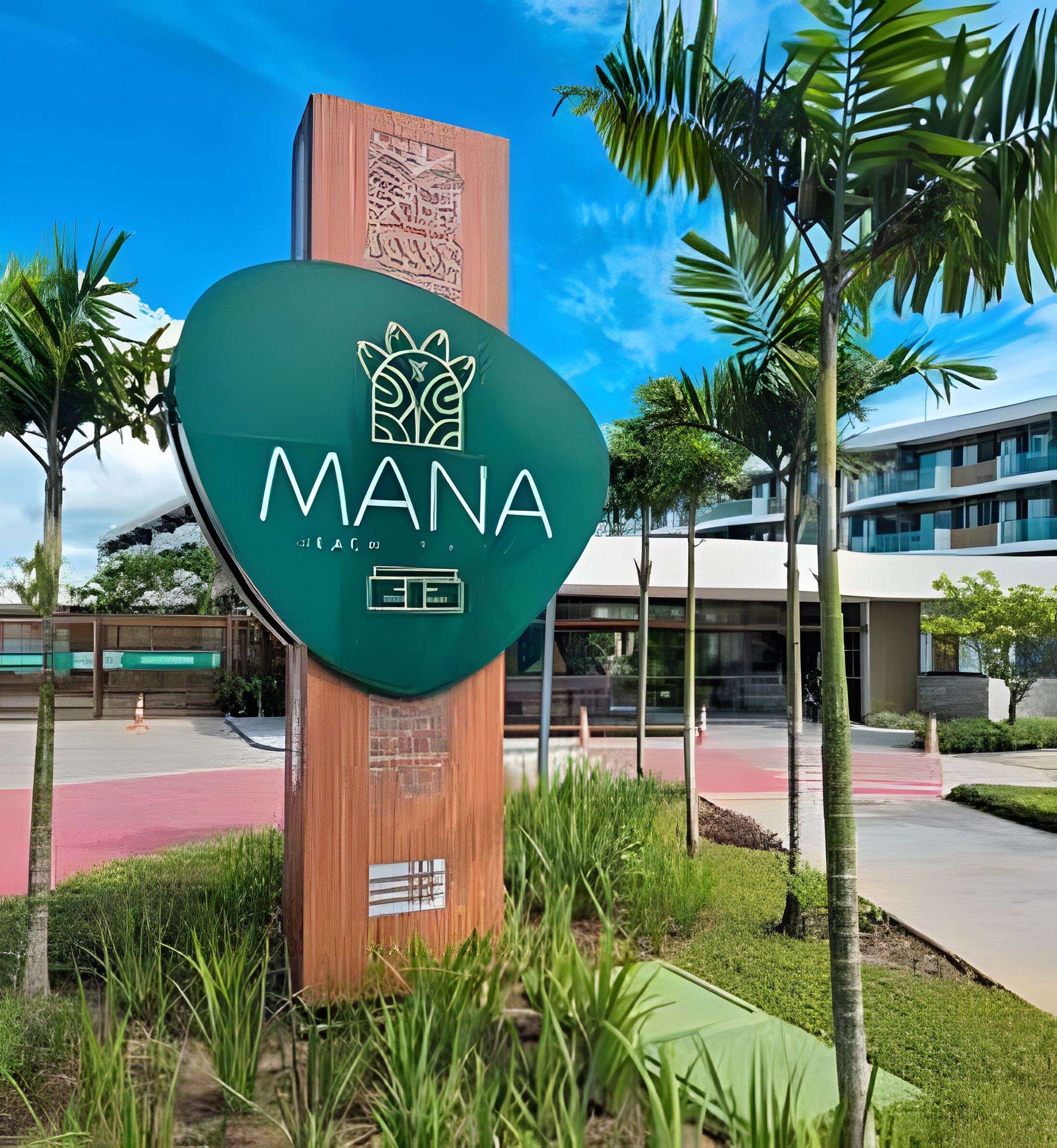 A sign with the word 'MANA' and a logo, surrounded by greenery and a modern building in the background.