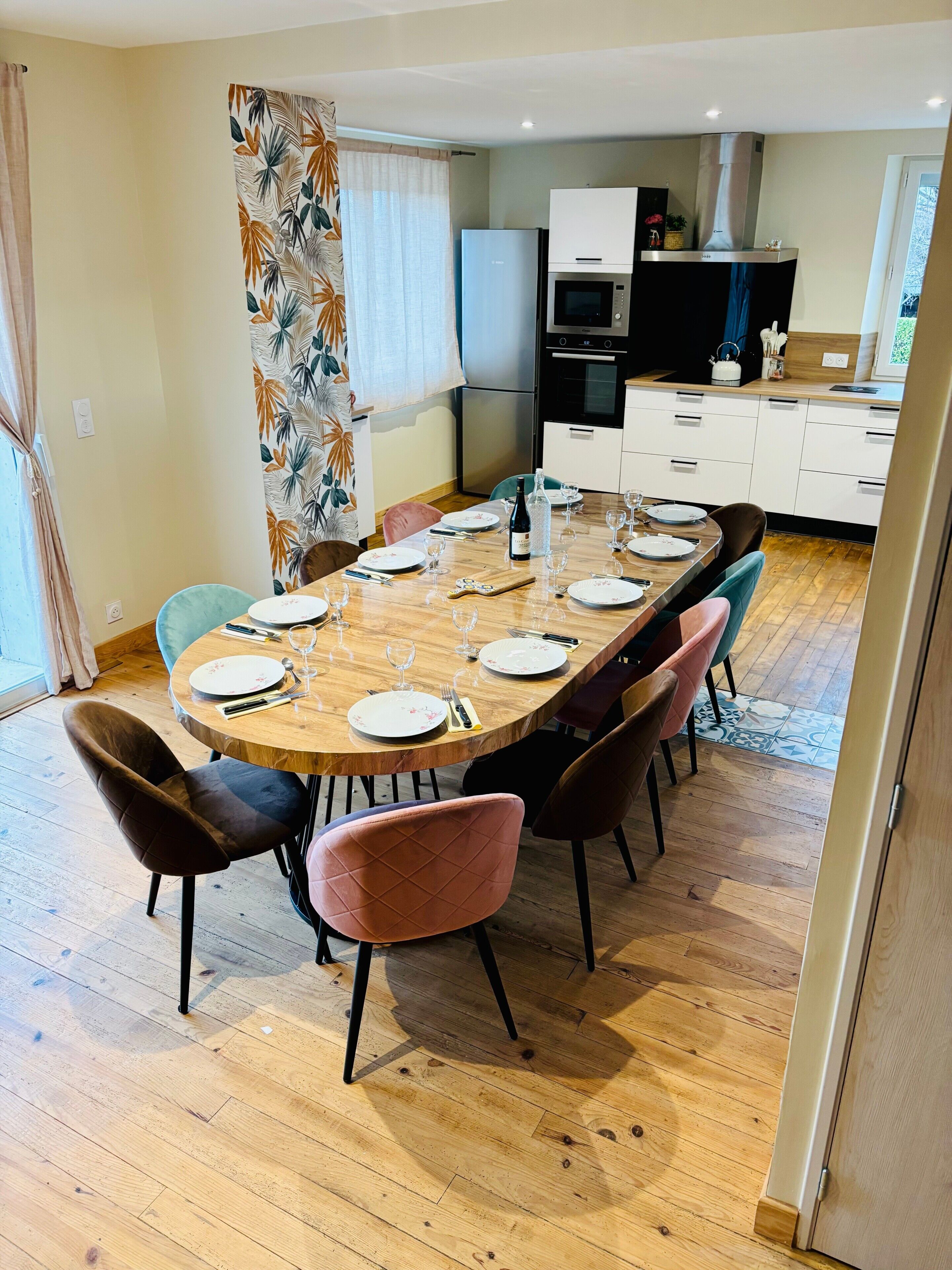 Un coin repas avec une table en bois dressée pour six personnes, entourée de chaises, dans une cuisine moderne dotée de meubles blancs et d’un four encastré.