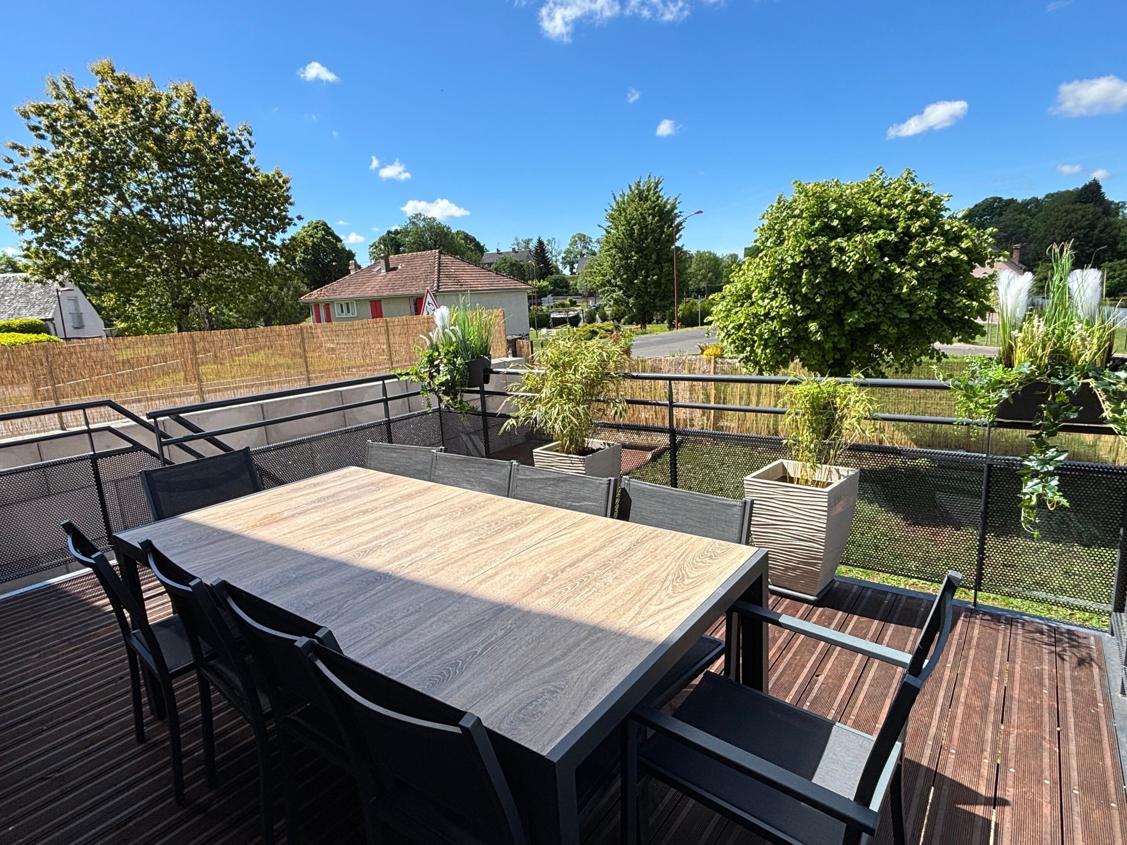 A wooden deck with a table and chairs, surrounded by a metal fence, overlooking a residential area with trees and houses.