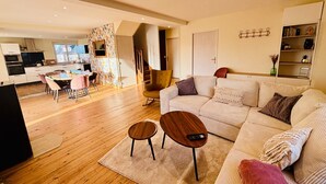 A modern living room with a sofa, two side tables, a dining area with chairs, a kitchen in the background, and a staircase leading to another level. - Chez Coco – 4★ Family Home, Jacuzzi & Games, Cantal (Mauriac)