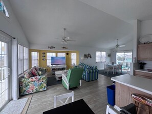 A spacious living room with a large TV, a green armchair, and a colorful couch. There is a kitchen area with a dining table and chairs. - Volare at the Sea 
3-bedroom townhome with Ocean View and Pool (North Topsail Beach)