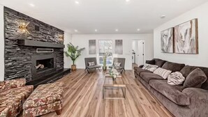 A modern living room with a stone fireplace, wooden flooring, a brown sofa, and two armchairs. - Walkout Bungalow with Pond Views (Delacour)