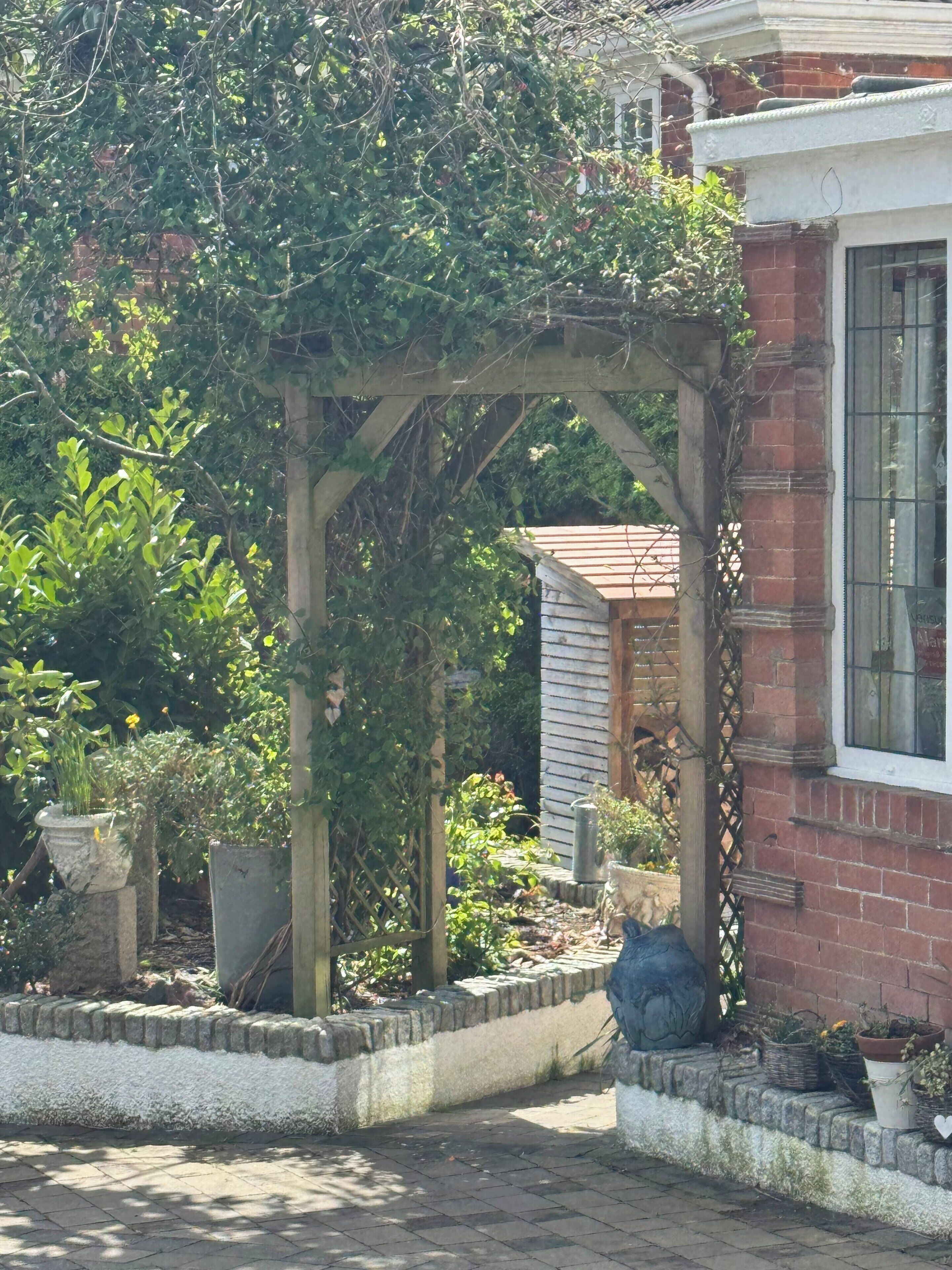 Uma área de jardim com pérgola de madeira, plantas em vasos e um edifício de tijolo com uma janela.