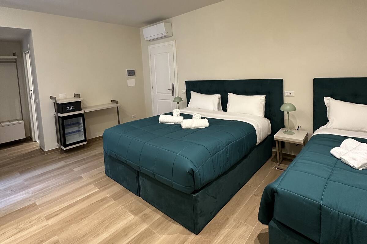 A hotel room with two beds, a small bedside table with a lamp, a wardrobe, and an air conditioning unit.