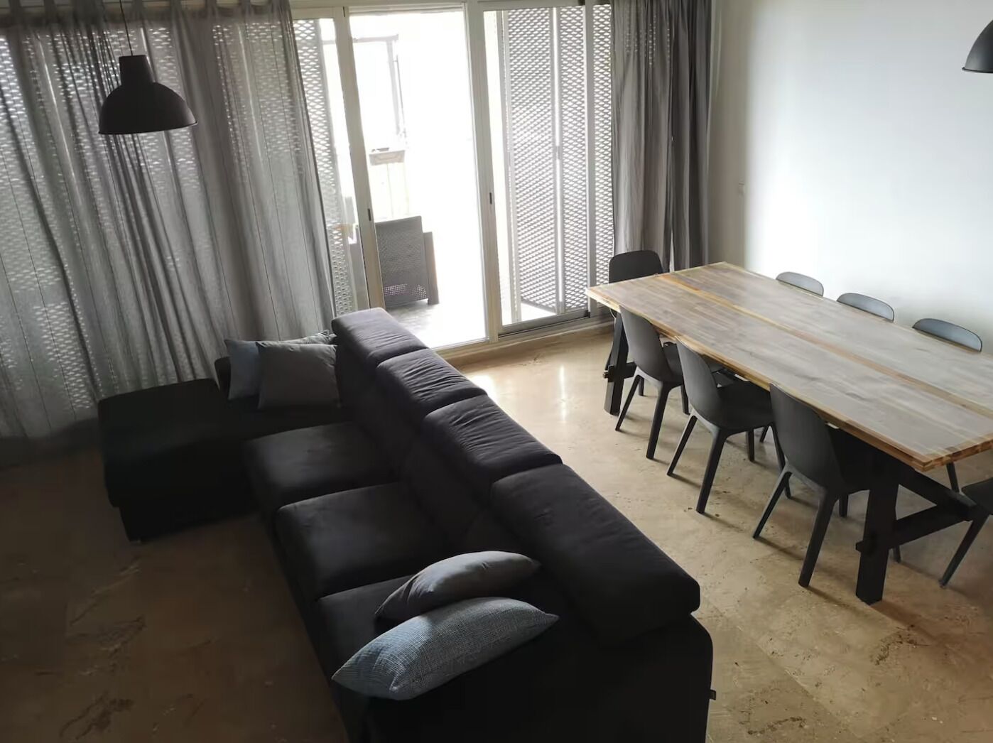 Un saló modern amb un sofà negre, una taula de dinar de fusta i un gran finestral amb cortines lleugeres.