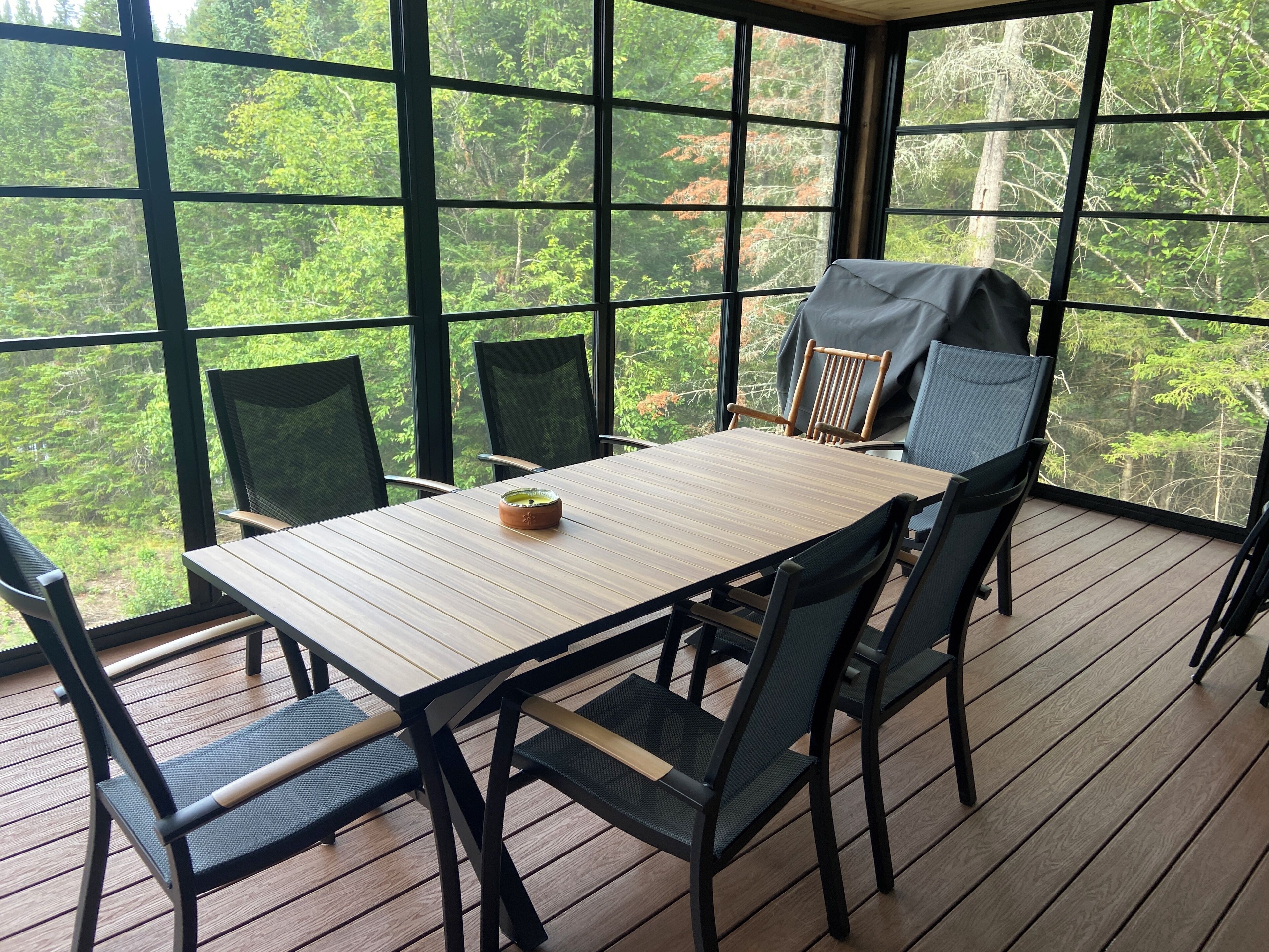 Área de refeições ao ar livre coberta, com mesa e cadeiras, cercada por grandes janelas que oferecem vista para uma área arborizada.