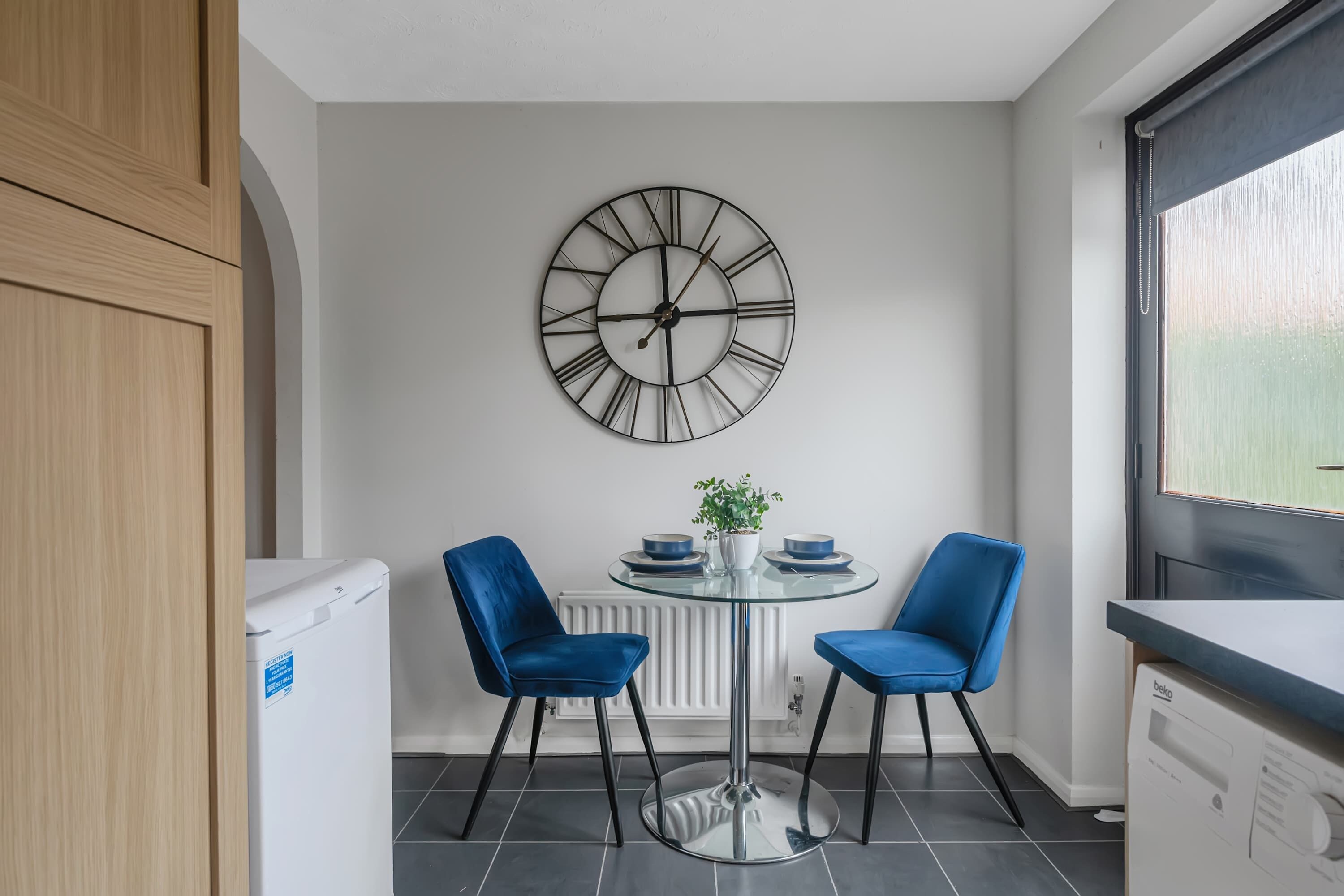 A modern kitchen with a round wall clock, two blue chairs, a small table, and a radiator.