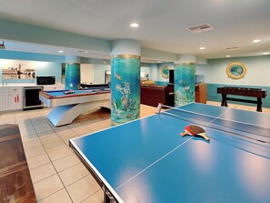 Une table de billard, une table de baby-foot et une table de ping-pong se trouvent dans une pièce aux murs de couleur turquoise et ornée de piliers décoratifs.