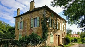 A rustic stone building with multiple windows and a door, surrounded by greenery and a dirt path.