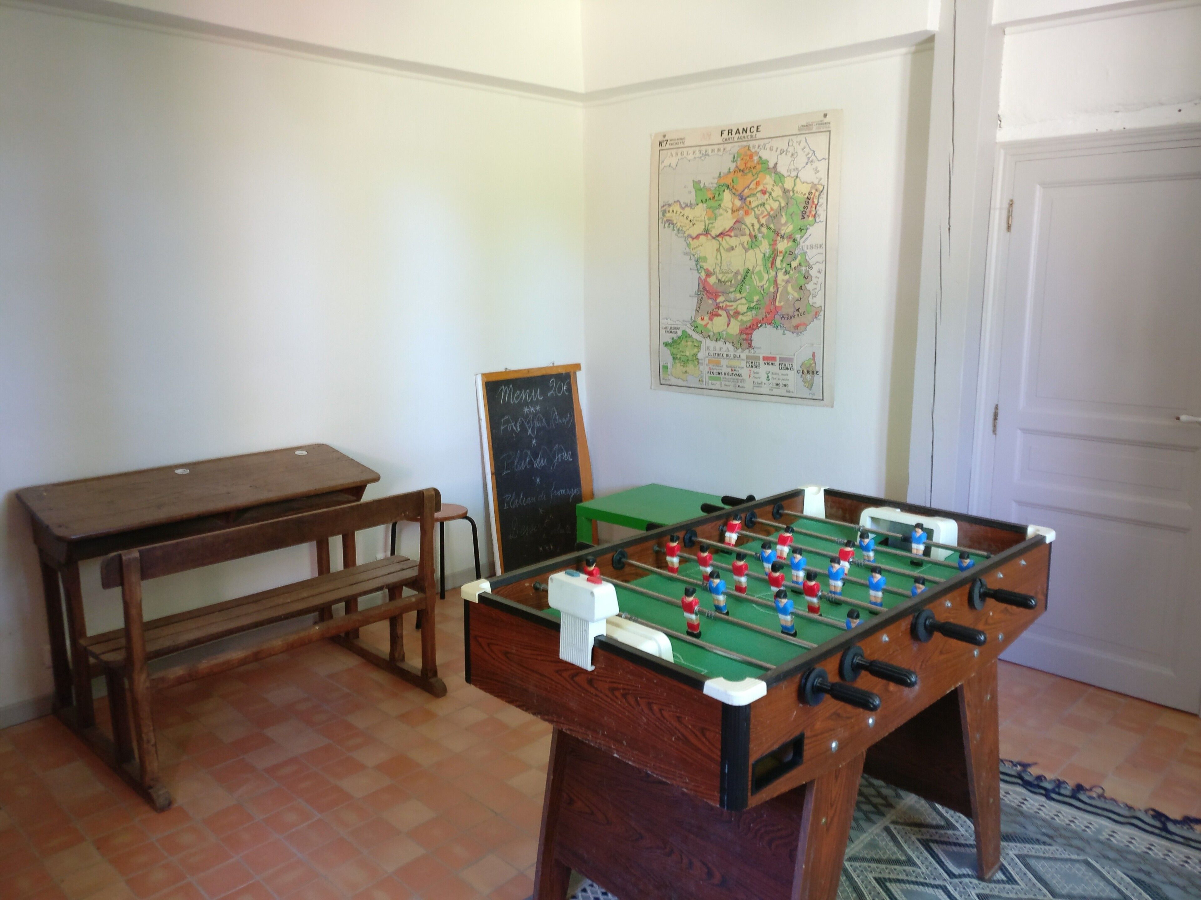 Une pièce avec une table de baby-foot, un banc en bois, un tableau noir servant de menu et une carte accrochée au mur.