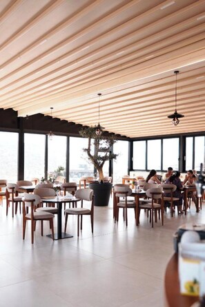 A modern restaurant with wooden ceiling slats, large windows, and a central potted plant. - Hotel Natica (Ksamil)