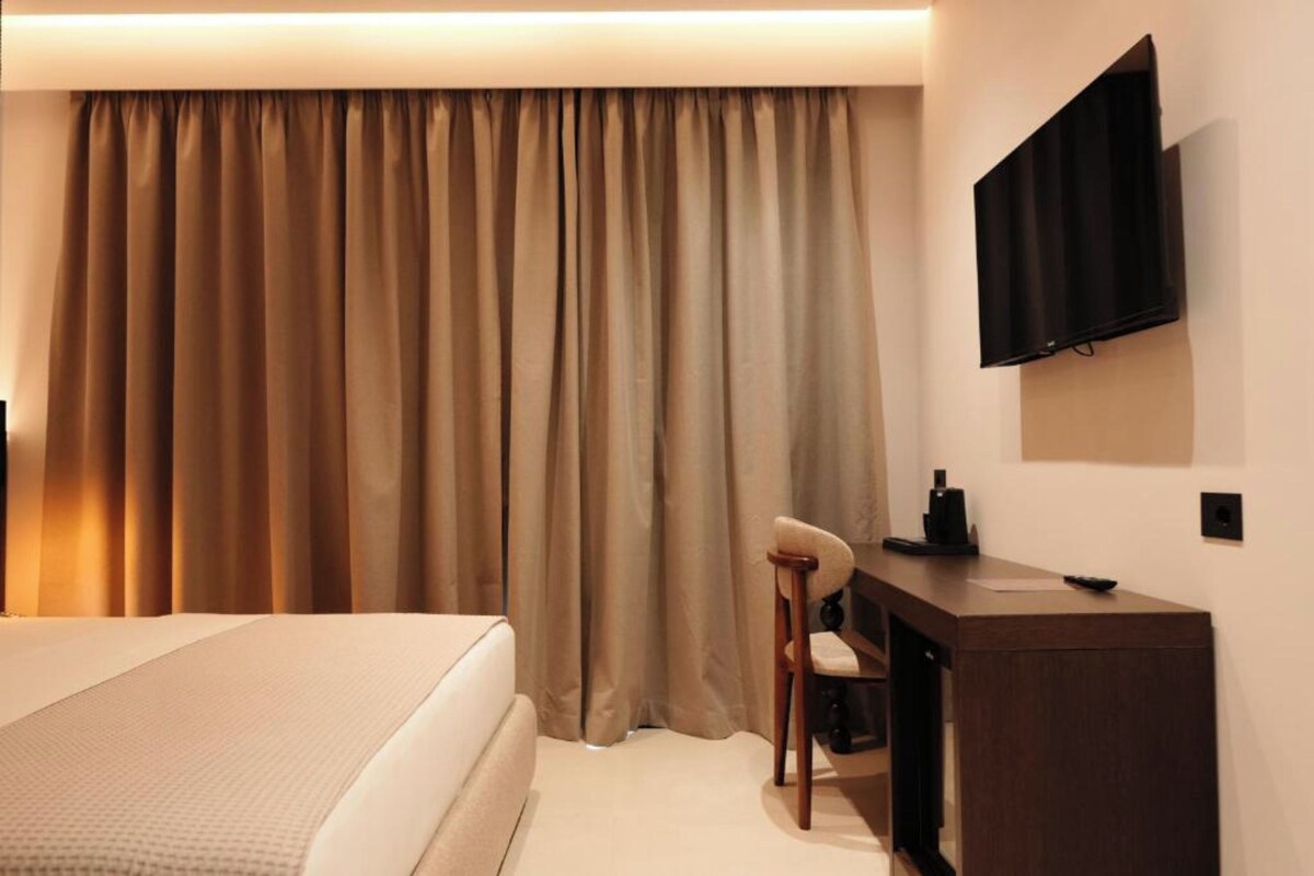 A hotel room with a bed, a desk with a chair, a television, and a large window with curtains.