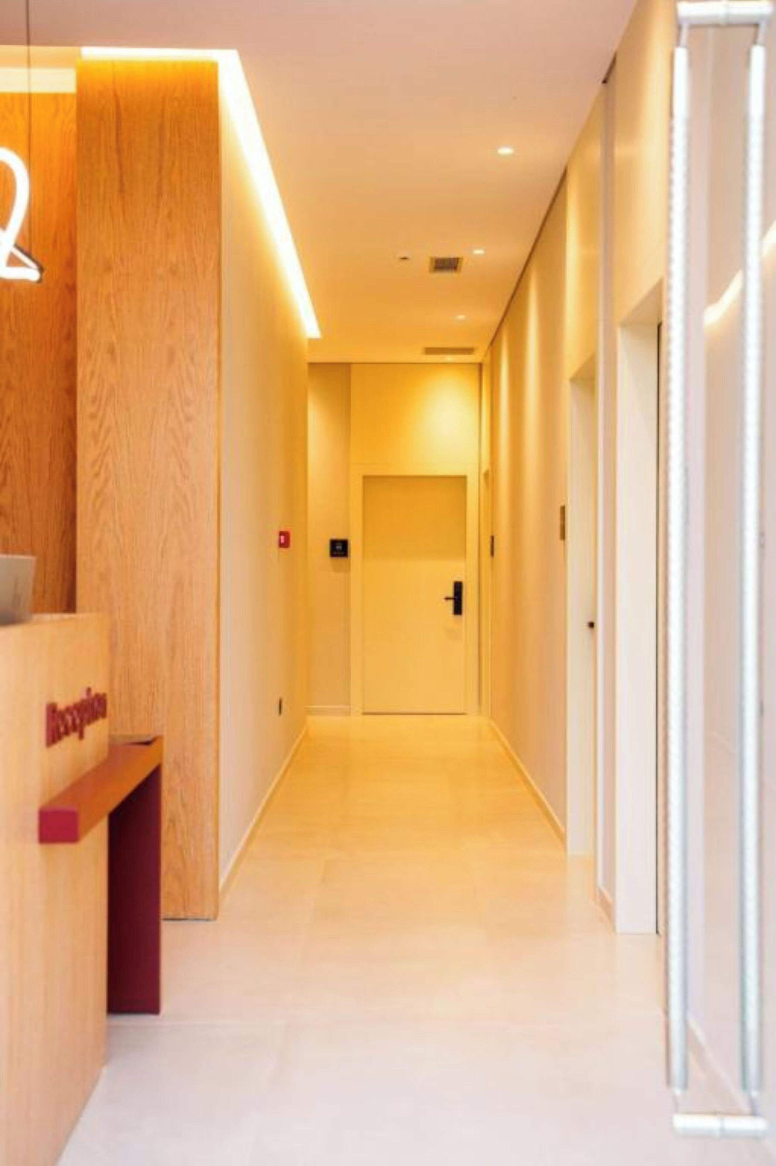 A modern hotel reception area with a wooden reception desk, a straight hallway leading to a door, and a clean, minimalist design.