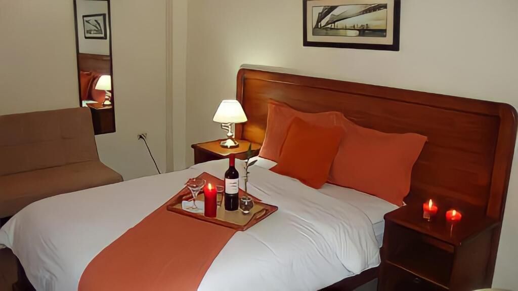 A hotel room with a large bed, a wooden headboard, a bedside table with a lamp, and a tray with a bottle and candles.