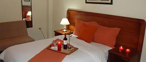 A hotel room with a large bed, a wooden headboard, a bedside table with a lamp, and a tray with a bottle and candles.