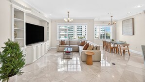A modern living room with a sectional sofa, a glass coffee table, a dining area with a glass table, and a large TV. - Ultra-Luxury Jupiter Condo!4 En-suite bedrooms! 100 Yards to Ocean! Game Room! (Jupiter)