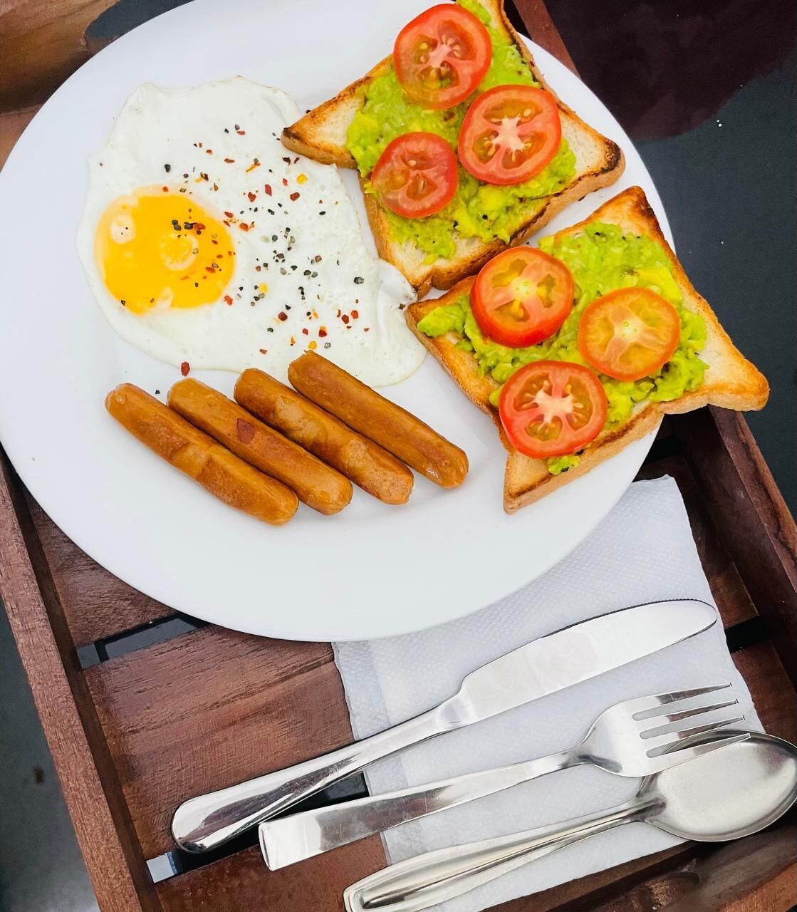 A plate with a fried egg, toast with avocado and tomato, and sausages.