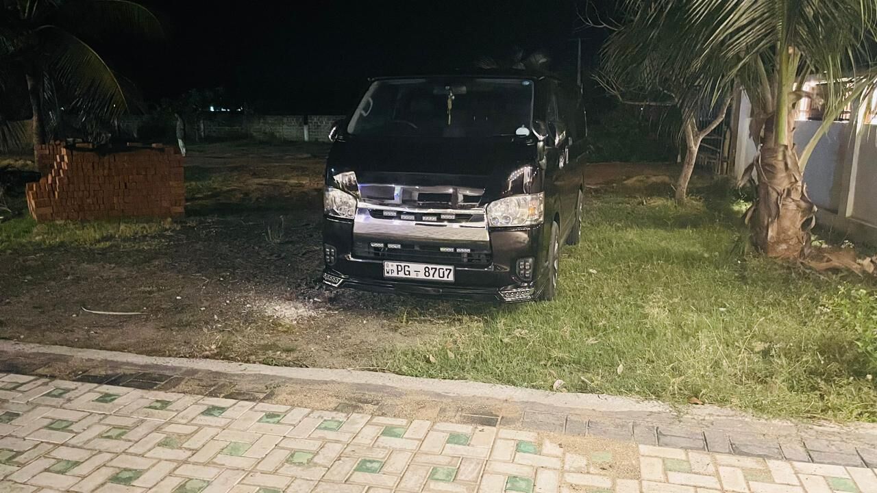 Um SUV preto estacionado numa área pavimentada, com grama e palmeiras ao fundo.