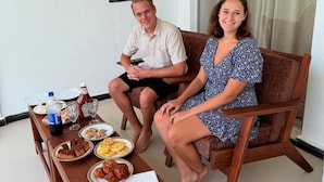 Ett par sitter på en träbänk i ett rum med en vit vägg och ett fönster med gardiner.