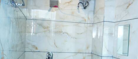 Un baño con ducha envuelta en vidrio, pared con azulejos de mármol, un lavamanos pequeño con dispensador de jabón y un taburete.