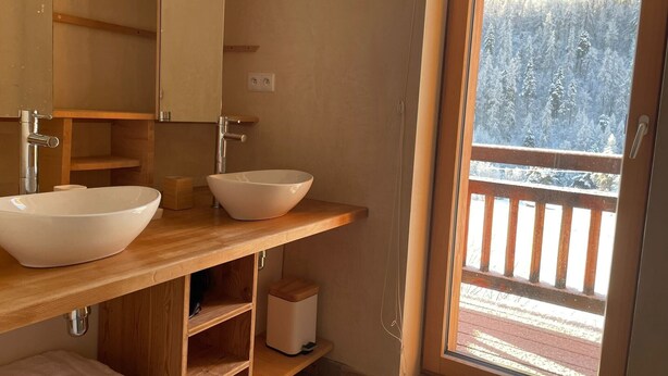 Ett badrum med två vita, fristående handfat, trähyllor, en spegel och utsikt över en snötäckt skog genom en glasdörr.