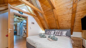 Un dormitorio con techo inclinado, una cama con almohadas y manta, una puerta de madera y un estante pequeño.