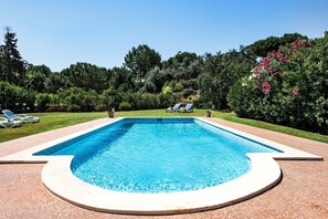 Une piscine rectangulaire bordée de blanc, entourée d’une terrasse en briques. On aperçoit des chaises longues et des plantes en pot à l’arrière-plan.