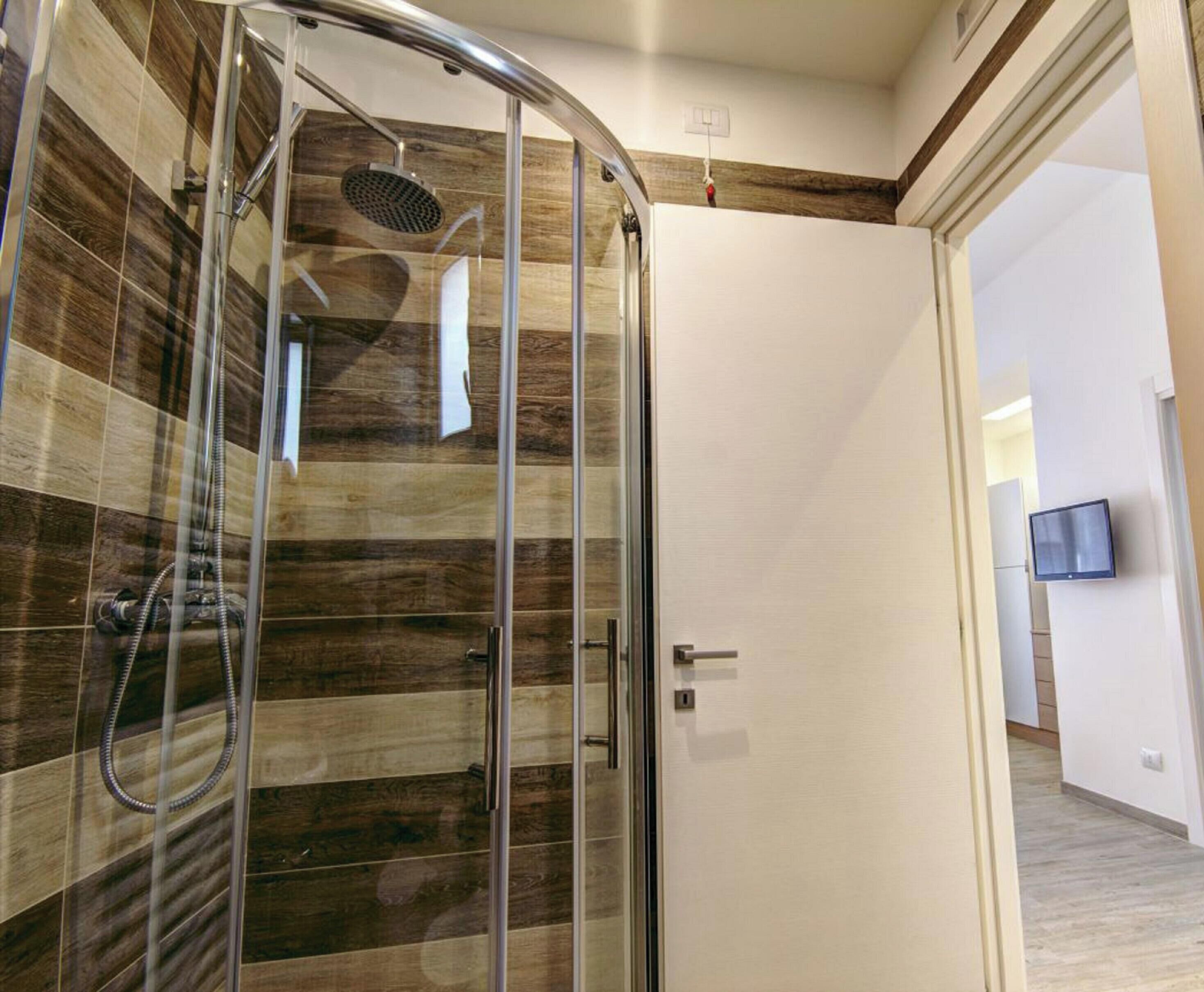 A modern bathroom with a glass shower enclosure, a white door, and a visible TV in the adjacent room.