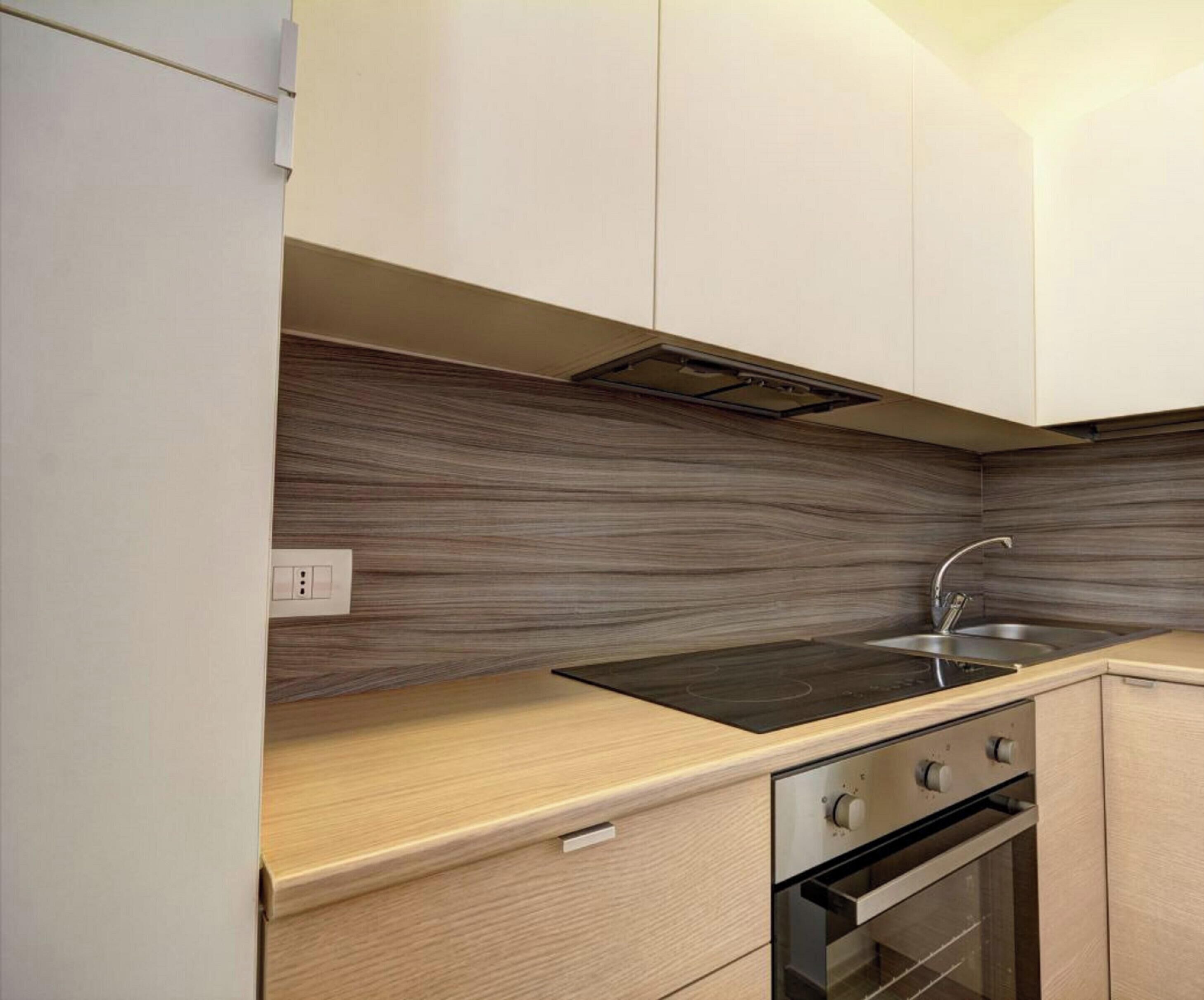 A modern kitchen with light wood cabinets, a black countertop, a stainless steel sink, and a built-in oven.