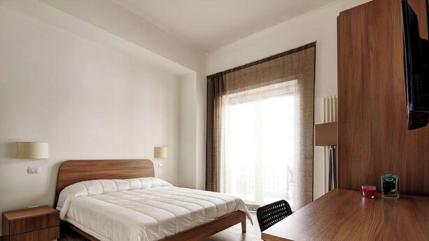 Habitación de hotel con una cama de madera, un escritorio, una silla, una mesita de noche, una lámpara y una ventana con cortinas.